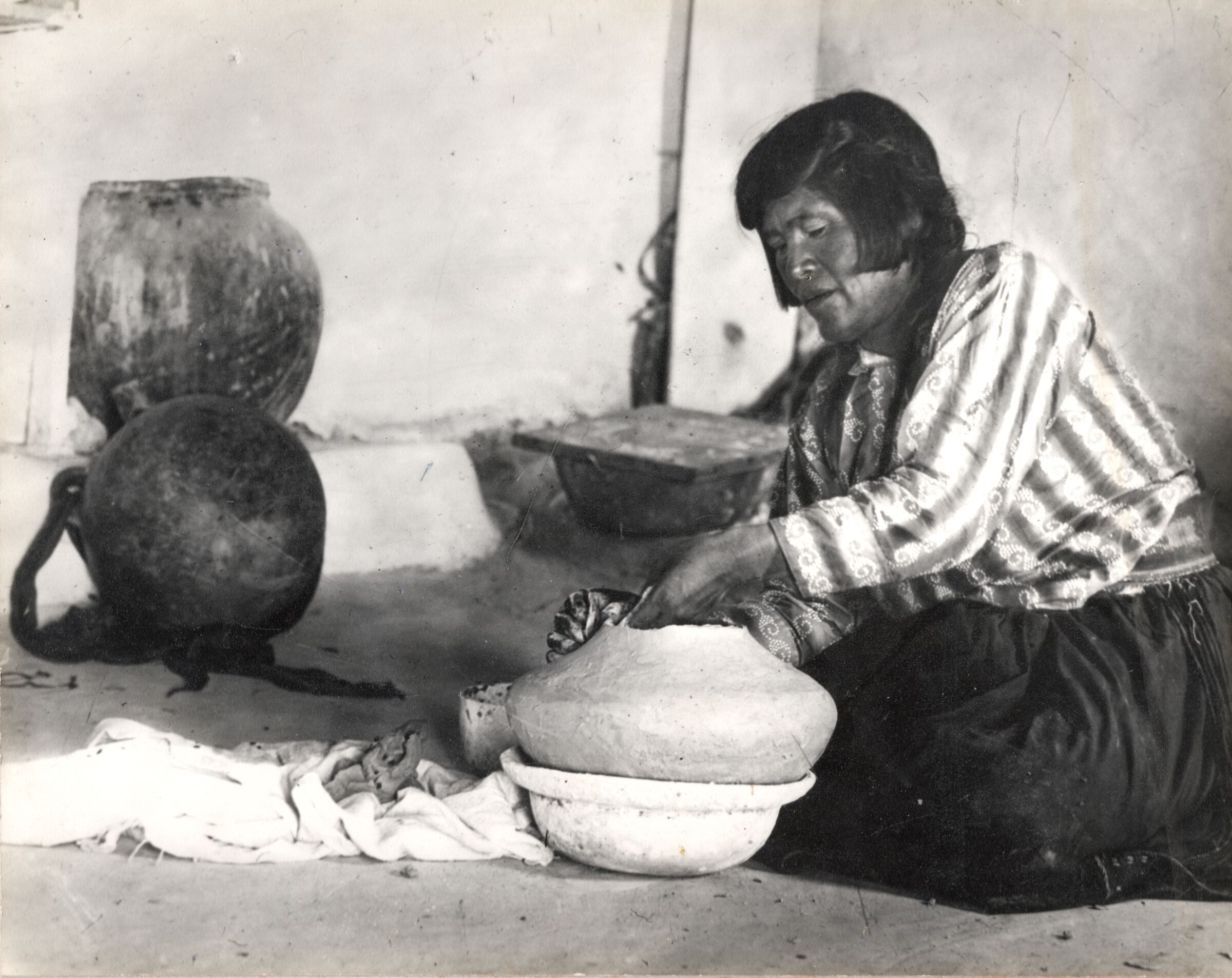 Historic photograph of Nampeyo seated on the floor, shaping a large pot, with storage jars behind her