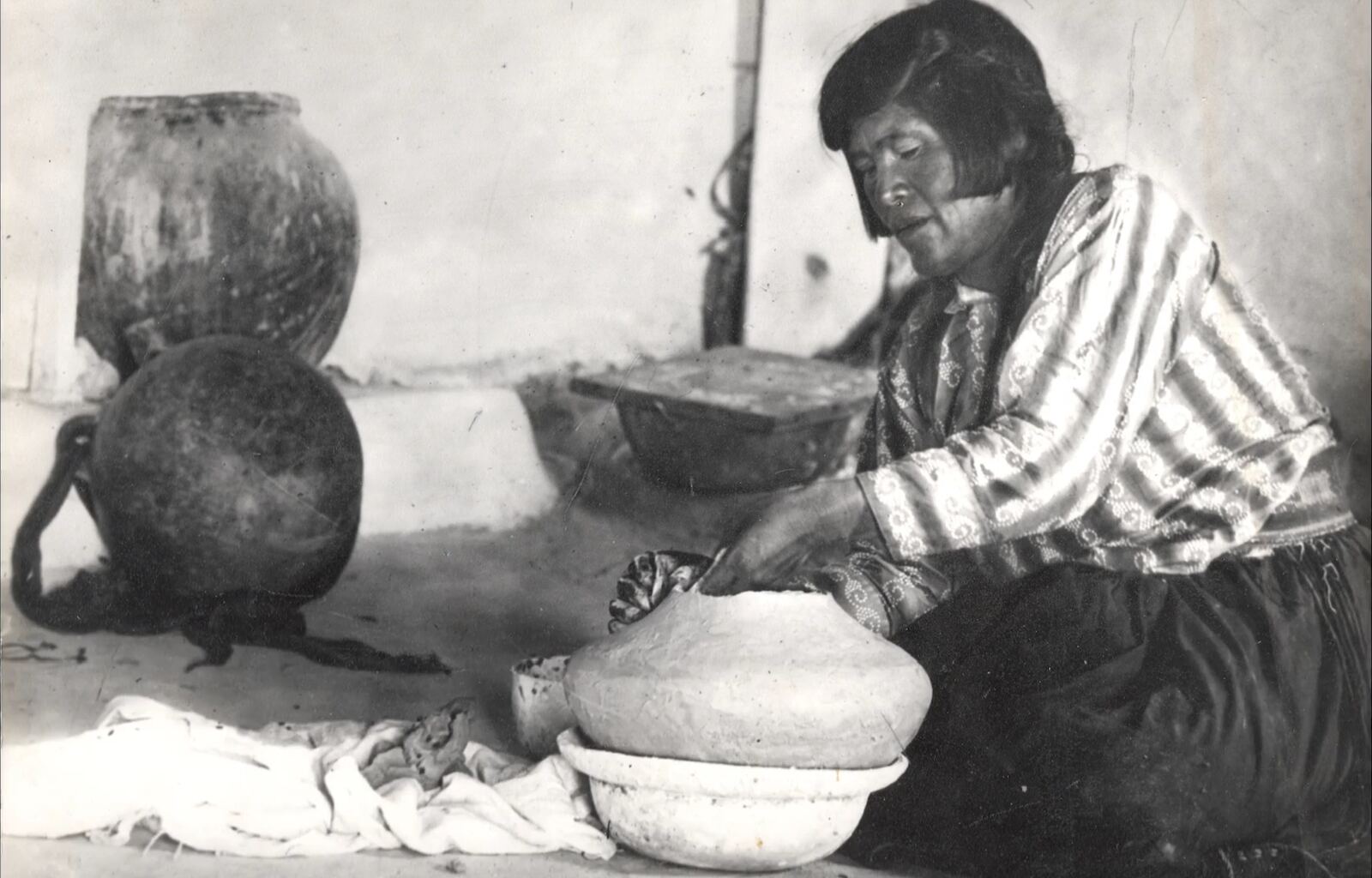Nampeyo seated on the floor of her home, shaping a large pot, with storage jars behind her
